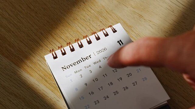Finger tapping dates on a small desk calendar for November 2026, angled view of the spiral bound page on a wooden desk crossed by warm sunlight and soft shadow, concept of autumn planning and countin