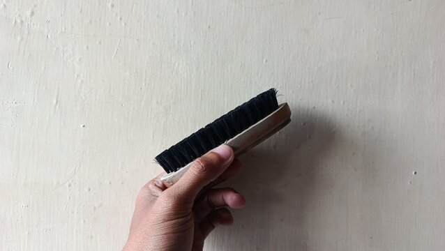 Hand holding a wooden shoe brush with black bristles against a plain wall background, concept for shoe care and cleaning services