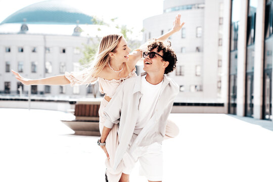 Young smiling beautiful woman and her handsome boyfriend in casual summer clothes. Happy cheerful family. Couple posing in street at sunny day. Female sits at his back. piggyback riding. Raise hands