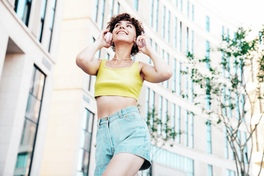 Young beautiful smiling hipster woman in trendy summer clothes. Carefree woman with curls hairstyle, posing in the street at sunny day. Positive model outdoors. Listens music at her headphones