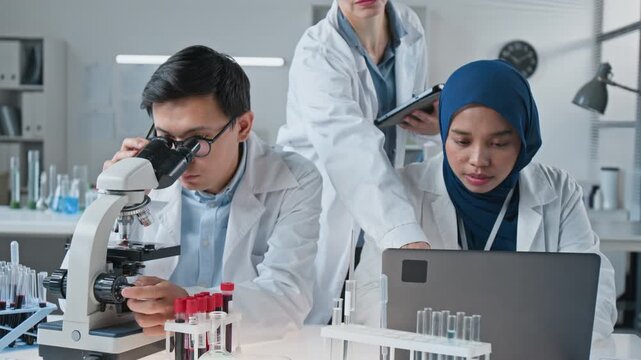 Diverse team of Asian man and Muslim woman analyzing samples using microscope and laptop in laboratory. Ideal for pharmaceutical development, medical research, and biotechnology campaigns