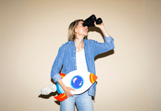 Smiling woman holds a paper rocket and looks through binoculars