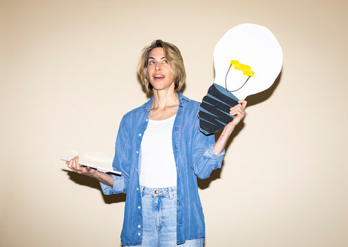 Inspired woman holds paper lightbulb and tablet indoors