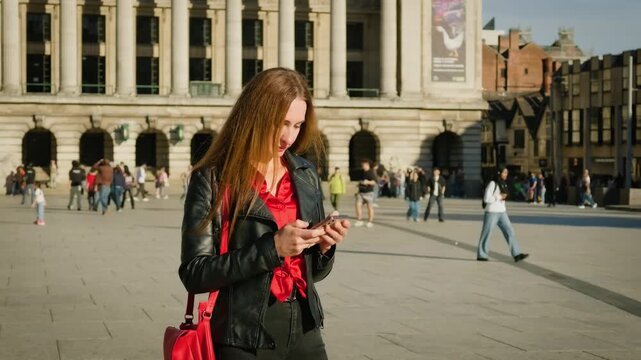 Text messaging in public square with historic architecture and pedestrians. Woman using smartphone outdoors in black jacket and red blouse. Mobile communication among city center crowd in daylight