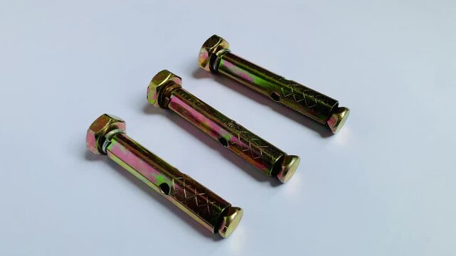 Real-time camera zoom in movement showing three yellow zinc plated sleeve anchor expansion bolts arranged on a clean white surface. High angle macro close up 4K video for concrete fixing and hardware 