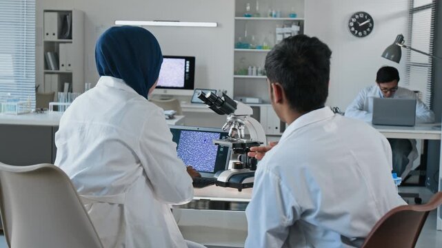 Asian young adult female in hijab and male colleague examining digital microscope in laboratory, rear view. Perfect for biotechnology research and healthcare innovation marketing