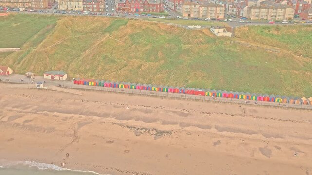 Colourful beach huts line the Lower Promenade beside Saltburn Beach, below the grassy coastal slope at Saltburn-by-the-Sea, Redcar and Cleveland, England, drone push in with descent.