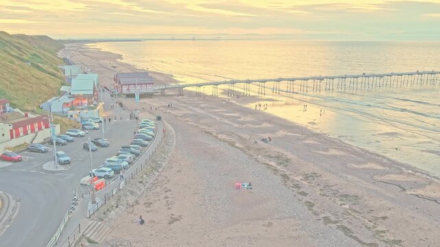 Saltburn Pier extends over the sandy foreshore beside the Lower Promenade, with beach facilities, coastal slopes and the North Sea visible at Saltburn-by-the-Sea, England, drone ascent.