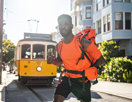 A focused Black man with a vibrant orange backpack jogs energetically through a historic city street, his determined expression and motion embodying urban adventure, active lifestyle, and resilience.