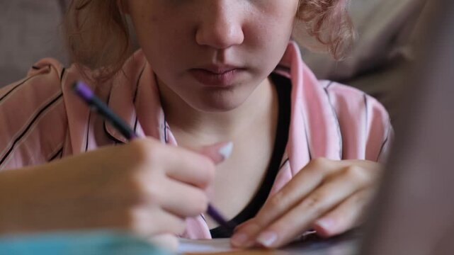 Close-up of a preteen girl in pajamas intensely shading a picture with a pencil at a small table, focused on her drawing hobby during free time, with children's art supplies, creative hobbies