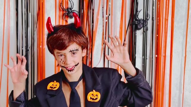 Medium close‑up of a child in a black jacket and tie with orange jack‑o&rsquo;‑lantern pins. The kid wears a red devil horns headband and stitched Halloween face paint, looks at the camera, raises hands lik