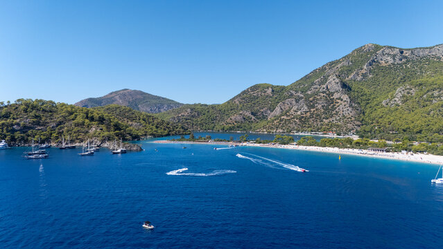 Aerial view of the famous Blue Lagoon in Oludeniz, featuring turquoise waters, sandy beach, surrounding pine-covered mountains, and boats anchored in the bay in Oludeniz, Mugla, Turkiye.