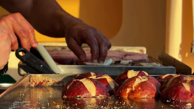 Mini burgers with pretzel buns on a hot griddle at a street food market, cook's hands flipping beef patties in warm golden sunset light