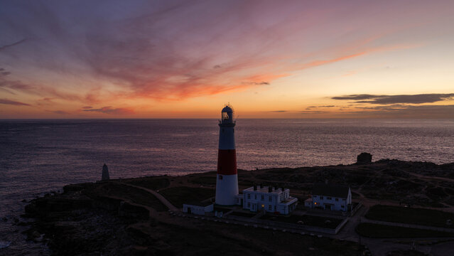 Aerial view of Portland Bill Lighthouse standing on the rocky coastline at sunset with a vibrant orange and purple sky over the English Channel in Portland, England, United Kingdom.