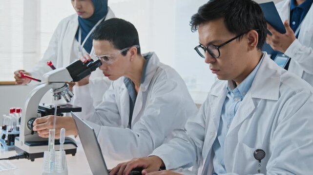 Caucasian female scientist and Asian male researcher analyzing clinical samples using microscope and laptop. Clip ideal for medical technology, pharmacy, biotechnology and science education projects