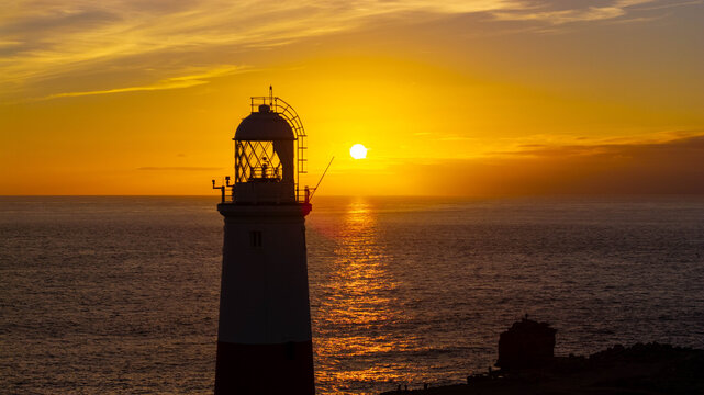 Aerial view of Portland Bill Lighthouse at sunset with the sun low over the sea casting a golden glow on the water and the lighthouse structure in Portland, England, United Kingdom.