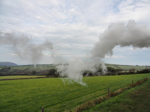 White steam and smoke billow across green rolling fields from Lynton and Barnstaple Railway heritage train in the scenic Exmoor countryside, Devon, UK