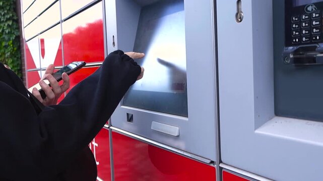 Young person using a modern self service parcel locker, entering a code on the touchscreen with a mobile phone in hand to retrieve a package from an automated post terminal outdoors