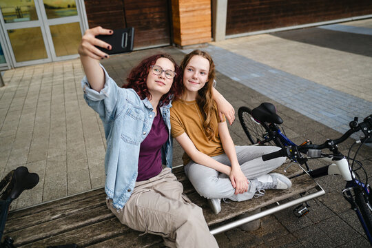 Teenagers sit on a bench outdoors taking a selfie with a smartphone