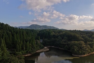 Fototapeta premium ドローンによる俯瞰空撮：青空と白い雲が広がる新緑の深い山林と静かな湖の水面を臨む日本の豊かな自然風景バックグラウンド（環境保全・SDGsイメージ・観光素材）