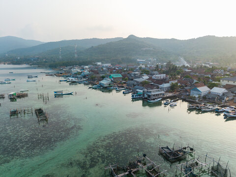 Aerial view of Karimunjawa Village with wooden boats, fish farming cages in clear turquoise water, and coastal houses nestled against green hills in Karimunjawa, Central Java, Indonesia.