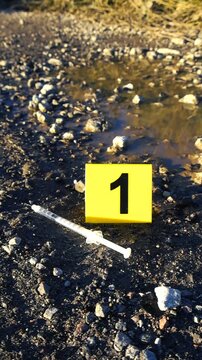 Crime scene investigation with a yellow evidence marker next to a used syringe on a muddy, gravel road during a sunny day. Forensic evidence of drug abuse or a murder is being examined