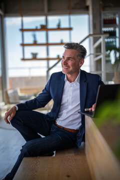 Smiling businessman sits on staircase with laptop indoors