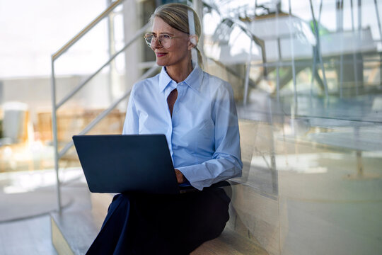 Smiling businesswoman uses laptop on office staircase in Germany