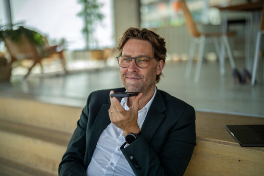 Smiling businessman records a voice message on his smartphone indoors