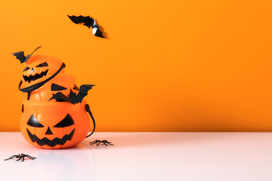 Halloween jack-o-lantern pumpkin buckets stacked with paper bats and plastic spiders on white table. Spooky October holiday background with flying bats and copy space on bright orange backdrop.