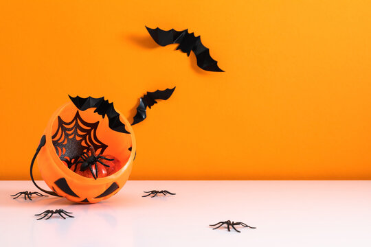 Halloween pumpkin bucket with spider web, decorative bat cuts and artificial spiders on table. Spooky October holiday background with flying paper bats and copy space on bright orange backdrop.