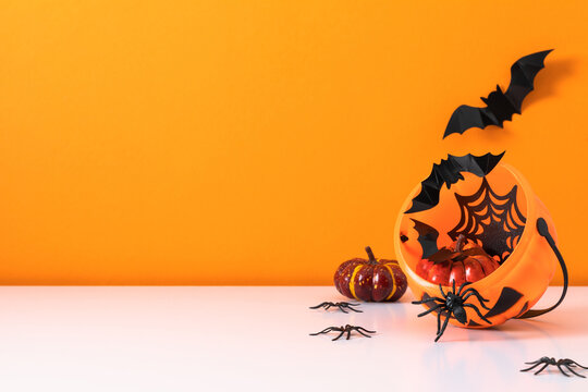 Halloween pumpkin bucket filled with paper bats, spider web and decorative pumpkins. Spooky holiday background with plastic spiders, flying bats and copy space on bright orange backdrop
