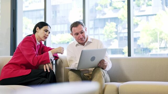 Two professionals sit on a sofa in a bright office, smiling and reviewing a laptop together during a collaborative meeting by large windows.