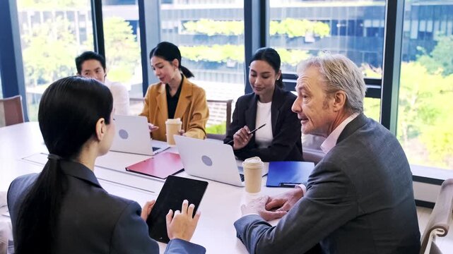 Business team meeting in a modern office, laptops and coffee on the table as colleagues discuss strategy by large windows with a city view.