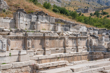 Large stone blocks and low walls spread across a dry hillside at Sagalassos. A carved facade bears shallow human reliefs beside an arched detail © Ensar