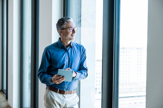 A businessman holds a tablet and looks out the office window