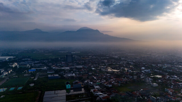 Aerial view of the Gedebage district featuring dense residential rooftops, misty mountains in the background, and soft sunset light in Bandung, West Java, Indonesia.