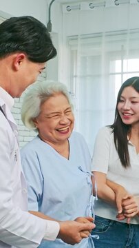 Happy elderly woman smiles with a doctor and younger family member in a hospital room, showing healthcare, recovery, support, caregiving, family, trust, joy and compassion.