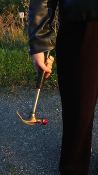 Murderer's hand holding a bloody hammer after committing a crime on a gravel road during a warm sunset. The killer is standing on the crime scene, a scary and disturbing concept
