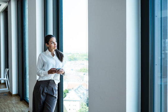 Thoughtful businesswoman by office window with smartphone