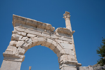 A weathered stone arch dominates the ruins at Sagalassos. Fluted columns and damaged capitals rise above sparse foliage beneath deep blue sky © Ensar