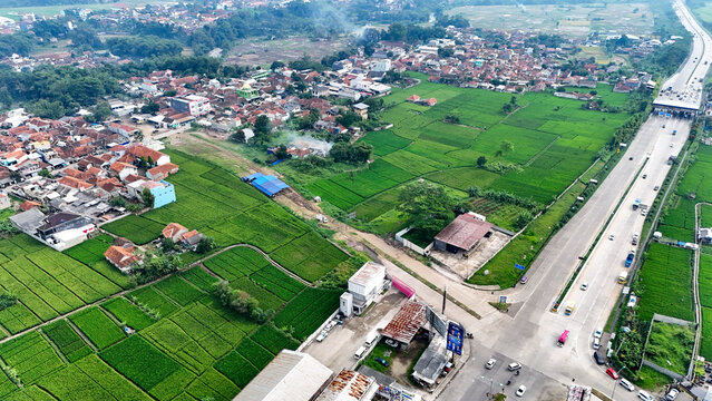 Aerial view of the Soreang area, featuring vibrant green rice fields adjacent to residential neighborhoods and a busy highway in Bandung, West Java, Indonesia.