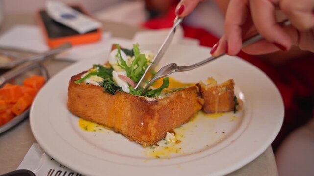 Two hands with red nails cut a thick golden brioche toast topped with fresh arugula and egg using fork and knife on a white plate, filmed in 4x slow motion 4K