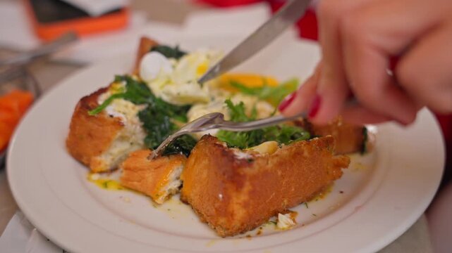 Womans hand with red nails cuts golden toasted bread topped with sauteed spinach poached egg and creamy yogurt on a white plate using fork and knife, filmed in 4x slow motion 4K