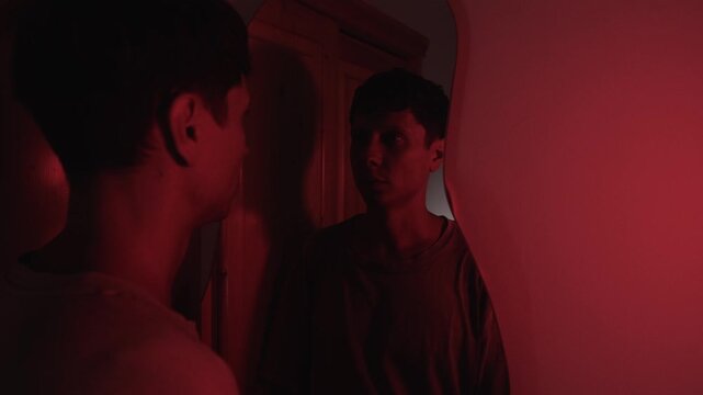 Man lowering his head before looking at his reflection and sighing, expressing emotional exhaustion, resignation and a difficult struggle with self acceptance in red low key light