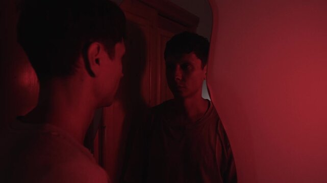 Man raising his head to look at his reflection, turning away before looking back again, expressing self doubt, insecurity and emotional conflict with his own image in red low key light