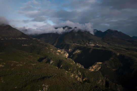 Aerial view of the rugged mountainous landscape of the Peloponnese, featuring deep valleys, dense green vegetation, and dramatic clouds clinging to the peaks, Peloponnese, Greece.