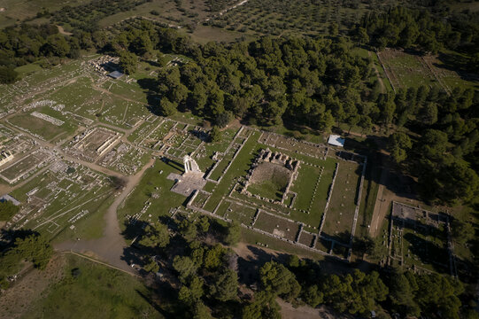 Aerial view of the ancient ruins of the Sanctuary of Asklepios at Epidaurus, featuring stone foundations and grassy courtyards surrounded by a dense pine forest in Peloponnese, Greece.