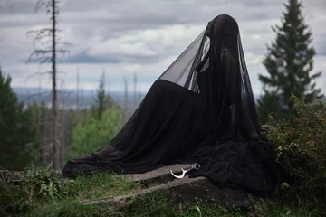 Woman sits beneath a stretched black veil on a rocky woodland ledge with a crescent prop resting beside her in a horizontal gothic portrait under a broad overcast sky © angel_nt