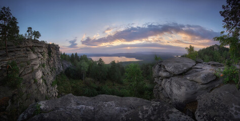 Rocky cliffs open toward a distant forest lake and low evening sun in a wide landscape photograph with golden clouds for rugged wilderness exploration and travel themes © angel_nt